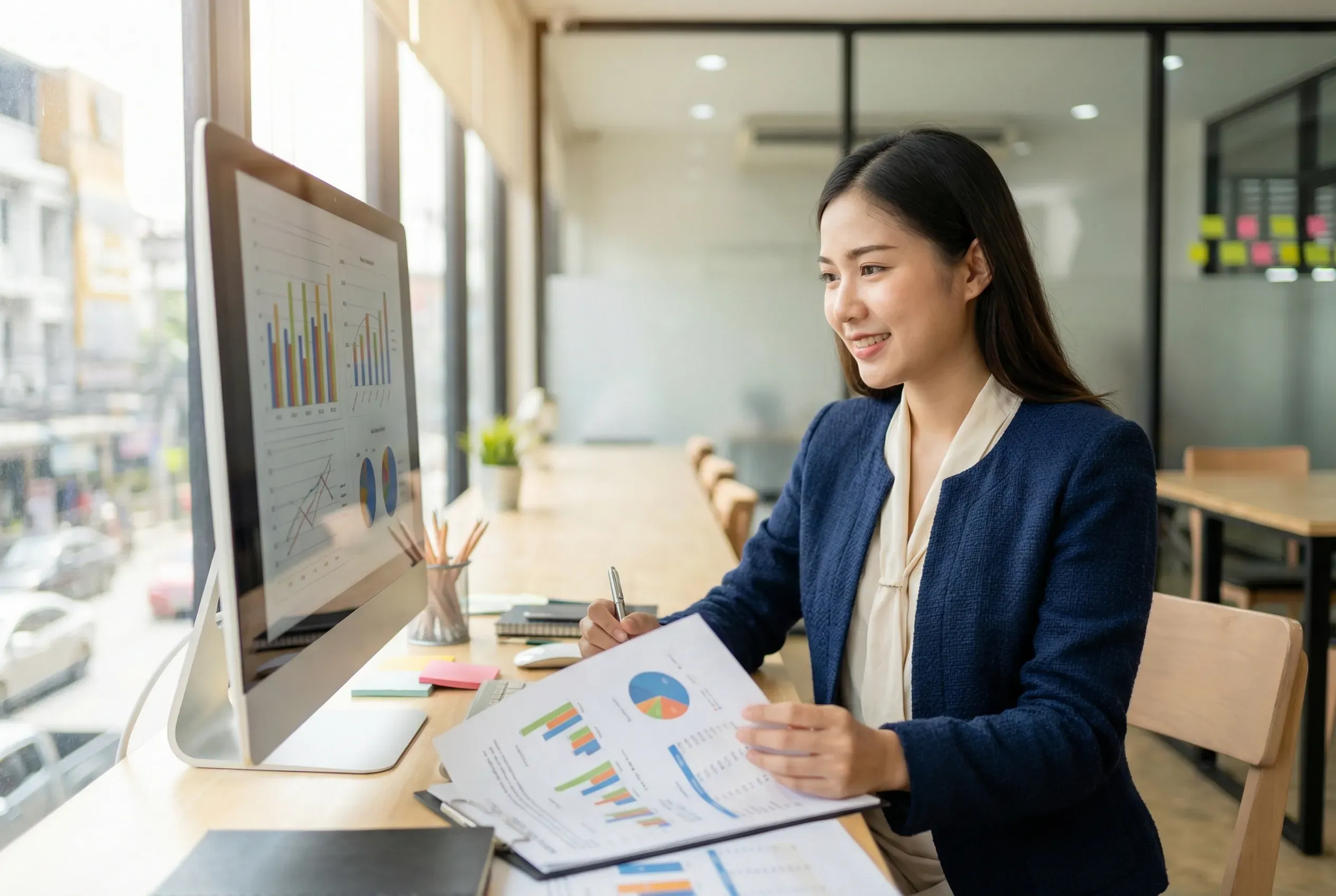 Business owner at her desk reviewing marketing budget allocation charts and growth reports on laptop and printed documents